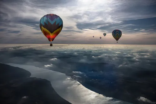 Horizontale luchtfotografie van luchtballonnen die boven een rivier en landschap vliegen tussen wolken