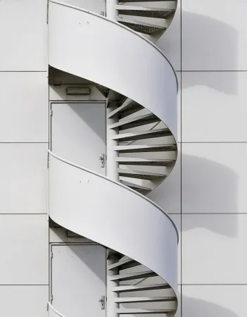 Verticale foto van een metalen brandtrap met spiraalvorm en geometrische schaduwen aan een witte gevel in Den Bosch