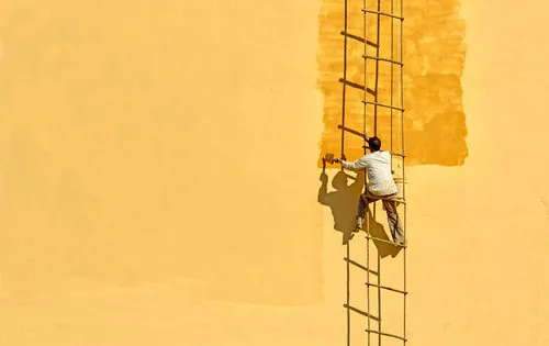 Horizontale foto van een man op een ladder die schildert in geel licht, met zachte schaduwen en een minimalistische uitstraling