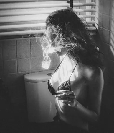Zwart-wit portret van een vrouw achter jaloezieën met rook in een badkamer, monochrome sfeer