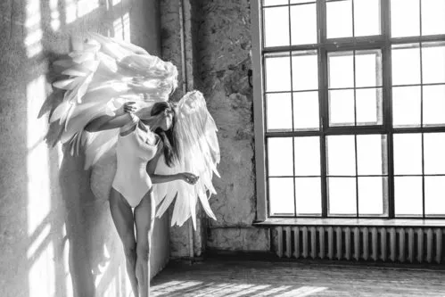 Zwart-wit horizontale foto van een dansende vrouw met engelenvleugels bij een raam in een studio