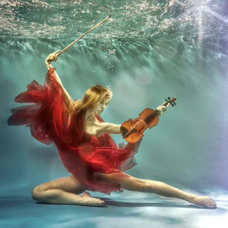 Vierkante fotografie van een vrouw die onder water viool speelt in rood licht, omgeven door bubbels en planten