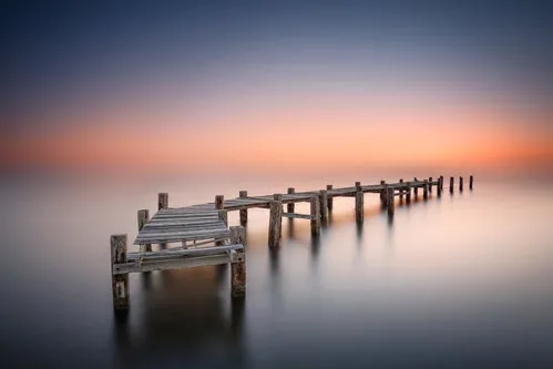 Horizontale foto van een oude houten pier bij zonsondergang met kalm melkachtig water en stille zee