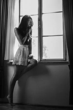 Zwart-wit verticale foto van een vrouw bij het raam met gordijnen, zachte lichtinval en sensuele uitstraling