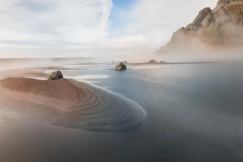 Horizontale fotografie van een mistige kustlijn met zand, rotsen en kalm water in een serene sfeer