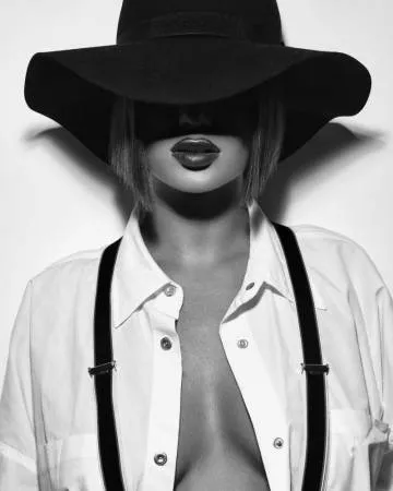 Verticaal zwart-wit portret van vrouw met hoed en bretels, hoog contrast en witte achtergrond